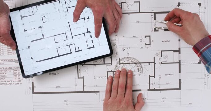 Two workers are working on a floor plan of a house, one taking measurements with a compass and protractor, the other moving an enlarged plan on an electronic tablet.