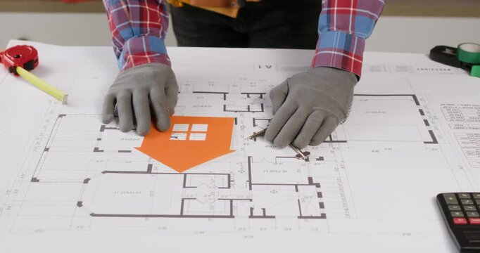 A worker in a plaid shirt places a house stencil and keys on a paper plan of the future house.