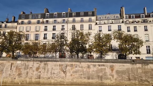 View from a cruising boat along the Seine river to the classic Parisian apartment buildings. Elegant Haussmann style facades and rows of trees with autumn leaves line the stone embankment