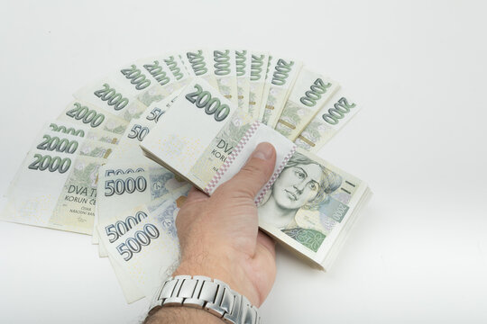 A hand holds Czech Koruna banknotes of 2000 and 5000 denominations, fanned out against a white background, photographed in the Highland region of the Czech Republic.