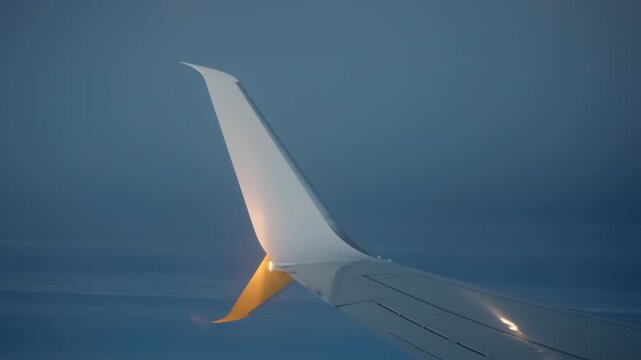 Close-up of aerodynamic aircraft winglet with glowing navigation light during evening flight. Sharklet wing tip cuts through dark blue twilight sky with illuminated position lamp at dusk.