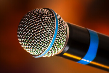 Close-up of a professional dynamic microphone head with a silver metal mesh grille. The audio equipment is isolated on a warm, blurred background, suggesting a stage, concert, or studio setting.