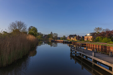 Peaceful morning by the canal in Bedum, Netherlands. Wooden pier, reeds by the shore and reflections of houses on the calm water surface