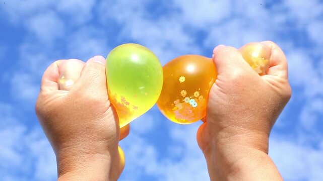 Two hands raise orange and yellow squishy gel balls toward a bright blue sky, squeezing and rotating them so glittery liquid shifts, then releasing as the spheres spring back in sunlight.