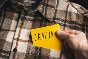 Yellow card with a handwritten inscription "Okazja", held in the hand against the background of a brown plaid shirt (selective focus)