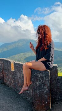 Red-haired girl wearing black t-shirt sits on the stone bastille. Picturesque mountains at backdrop. Vertical video.