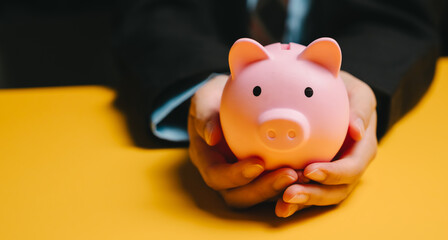 Pink piggy bank held by hands on yellow surface symbolizing saving money and financial security