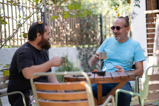 Men Relaxing With Coffee or Tea at Outdoor Table. Friends Enjoying Hot Drinks Outdoors in Garden. Casual Conversation Over Hot Drinks in the Garden. Men Sitting at Garden Table Drinking Hot Beverages.