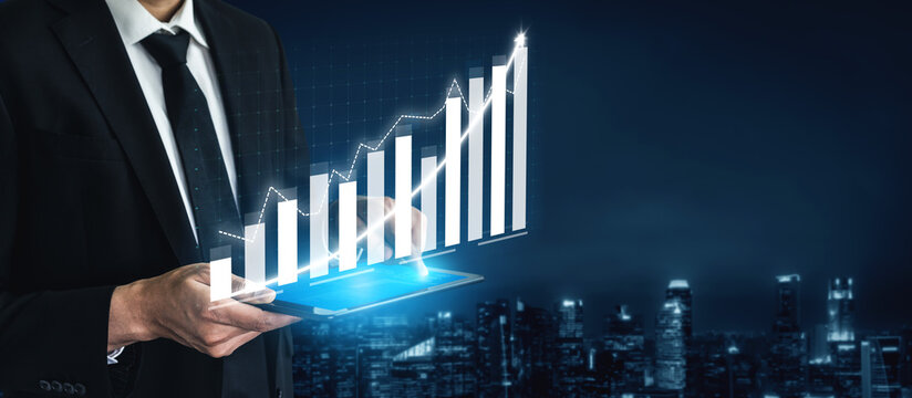 A businessman stands against a night city skyline, holding a tablet displaying a digital financial graph that symbolizes growth and success in business. Impute - Powered by Adobe