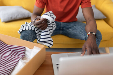 Young African man using laptop to manage online clothing return at home after delivery. E-commerce, technology and everyday life at home.