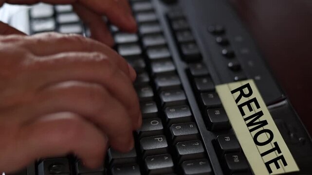 Un primer plano de un teclado de una computadora con las manos de una mujer adulta y una nota amarilla con la palabra remoto