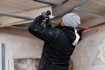 Worker using a cordless drill overhead, fastening a ceiling frame in an unfinished room. Winter hat...