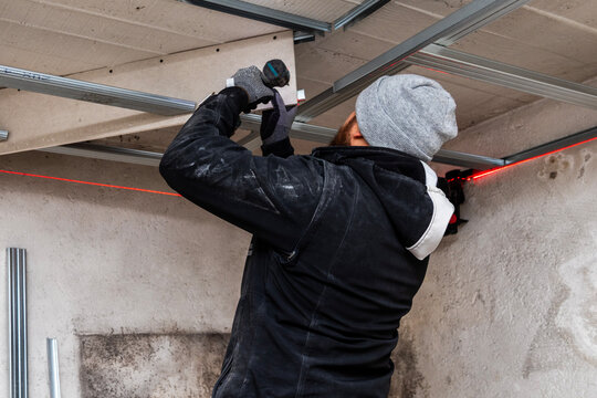 Worker using a cordless drill overhead, fastening a ceiling frame in an unfinished room. Winter hat and gloves, indoor renovation and construction maintenance concept. - Powered by Adobe