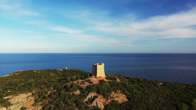 Aerial view of a historic stone tower overlooking the blue Mediterranean Sea on ile de Pinarello, Corsica, surrounded by lush greenery under a clear sky.