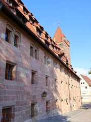 A sunlit historic building with pink stone, red-tiled roof, and a tall tower lines a quaint cobblestone street in old town, Nuremberg, Germany. Perfect for themes of heritage, architecture and travel