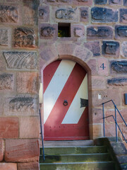 A historic stone arch frames a bold red door with diagonal white stripes, set into a textured masonry wall. The number four and metal railings hint at an old building entrance.