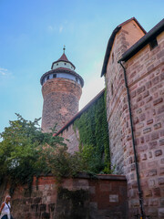 Ancient Stone Tower Rises Over Ivy Covered Wall Under Clear Blue Sky. Tall medieval tower stands beside an ivy-clad wall next to a brick building. Perfect for architecture, history and travel imagery.