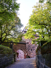 Tranquil medieval scene in Nuremberg, Germany, featuring rugged stone gate and arched passageway framed by lush green trees. Sunlight filters through leaves, casting dappled light on cobblestone path