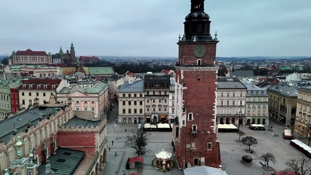 Low drone fly-by reveals the intricate clock face of Krakow's Town Hall Tower with Sukiennice and Wawel Castle visible in the distance.