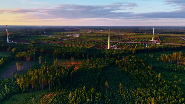 A wide aerial shot of a wind farm under construction at sunset, showing multiple tall turbine towers rising above a patchwork of forest and cleared land, developing infrastructure.