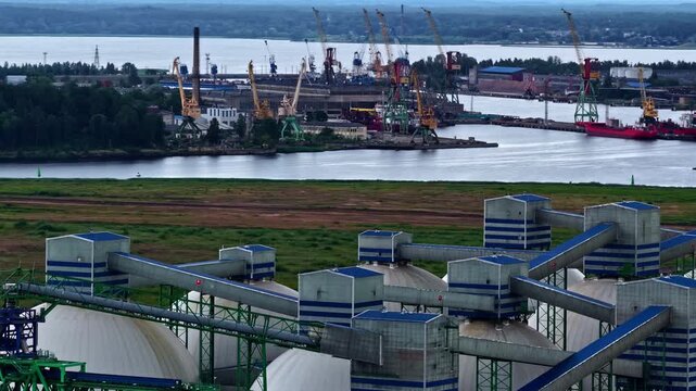 A busy industrial port stretches along the water, with cargo cranes, docks, and ships in the background, while large domed storage buildings and conveyor systems dominate the foreground.