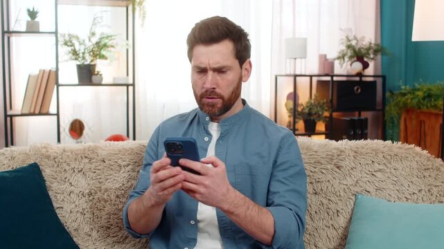 Middle-aged man at home feeling frustrated and nervous while texting on smartphone, holding head in distress. Nervous Caucasian guy sitting on sofa, anxiously waiting for an important message reply