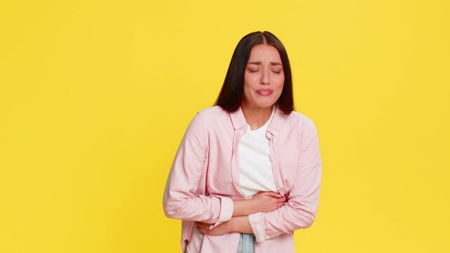 Young woman rubbing stomach bending forward with painful expression, clearly suffering from abdominal discomfort. Girl on yellow background shows visible symptoms of stomach pain and digestive issue.