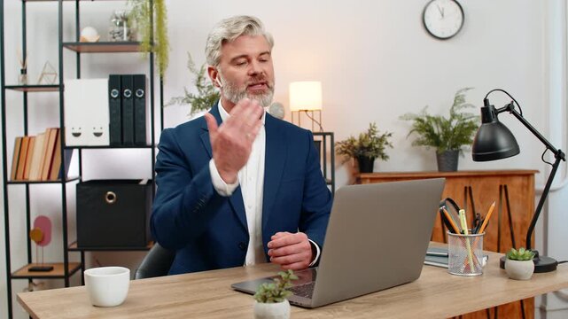 Middle-aged businessman at home office hosts webinar using earphones through laptop presenting topic. Freelancer guy at table answers audience questions sharing expertise guiding learners to success.