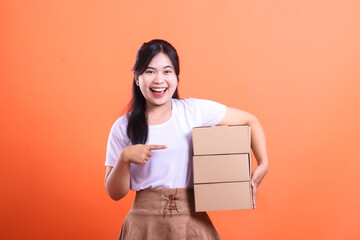 A smiling woman holds three cardboard boxes, and with her other hand, points at them, showcasing the delivery. isolated on orange background