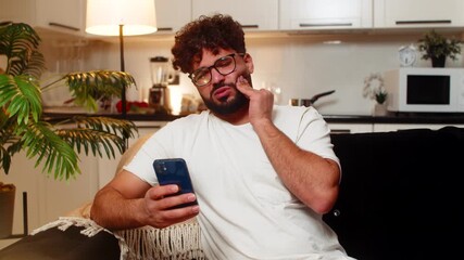 Arabian man at home feels tooth pain holds cheek opens smartphone search for dentist finds time. Egyptian guy on sofa reads reviews picks dentist sends message books visit for pain relief needs care.