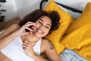 Young happy Latin American woman with curly hair smiling while making a phone call lying in bed with yellow pillows
