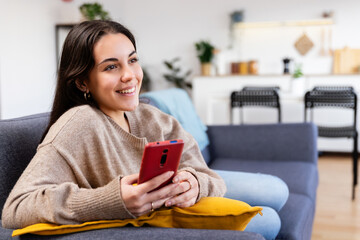 Young gen z woman relaxing on sofa using mobile phone at home, connecting online with friends and browse social media in a cozy living room. Technology lifestyle concept