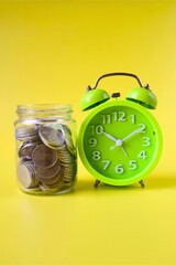 Green alarm clock and jar filled with coins on yellow background