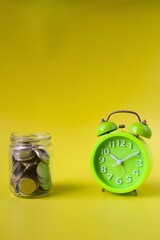 Green alarm clock and jar filled with coins on yellow background