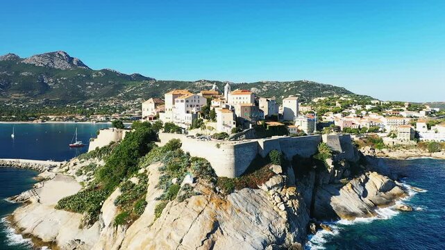 Aerial view of the historic citadel perched on a rocky coastline in Calvi, Corsica, with mountains and blue sea in the background under clear skies.