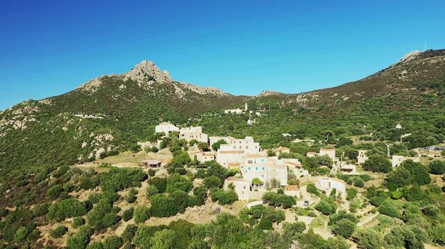 Aerial view of the picturesque hilltop village of Pigna surrounded by lush green hills and mountains under a clear blue sky in Corsica, France.
