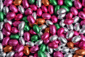Pink, green, orange, silver and bronze foil wrapped chocolate easter eggs, against a black background.