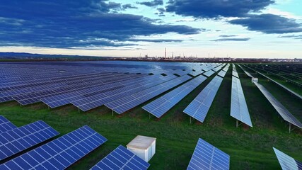 Rows of solar panels stretch across a green field, reflecting the soft glow of the setting sun. In the background, industrial structures rise against dramatic clouds, creating a striking contrast.