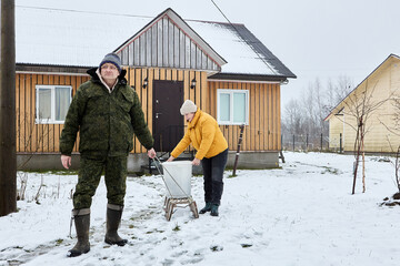 Man pulls sled by rope leaving rural house, woman holds garbage bin standing on it.
