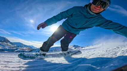 CLOSE UP, LENS FLARE, LOW ANGLE VIEW: Happy snowboarder carving on sunny ski slope. Smiling young man enjoys shredding at ski resort. Winter activity and authentic snowboard action in Austrian Alps. © helivideo