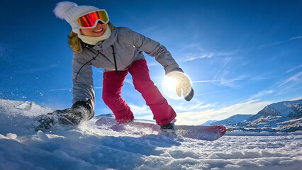 LENS FLARE, LOW ANGLE VIEW, CLOSE UP: Cheerful female snowboarder drags her hand and sprays snow while she carves across a sunlit slope. Winter fun and authentic snowboard action in Austrian Alps. © helivideo