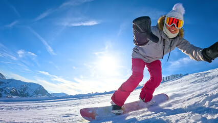 LENS FLARE, LOW ANGLE VIEW, CLOSE UP: Smiling female snowboarder carves across a sunny ski slope, reaching hands toward the camera. Fun snowboarding in snowy mountains of Turracher Hohe, Austria. © helivideo