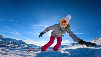 LENS FLARE, LOW ANGLE VIEW, CLOSE UP: Smiling female snowboarder carves past the camera on snowy piste at ski resort. Snow sprays up around her hand as she leans into turn under bright winter sun. © helivideo