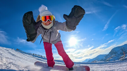 LENS FLARE, LOW ANGLE VIEW, CLOSE UP: Happy woman playfully leans towards camera with gloved arms outstretched while snowboarding, framed by blue winter sky and sunbeams over snowy Austrian mountains. © helivideo
