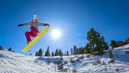 LOW ANGLE VIEW, LENS FLARE: Female snowboarder performs a stylish nose grab during a jump while riding through snowpark at ski resort on a sunny winter day. Fun winter days in snowy Austrian mountains © helivideo