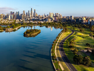 Obraz premium Morning aerial panorama of Melbourne's modern skyline reflecting in Albert Park Lake with vibrant green parklands and pathways.