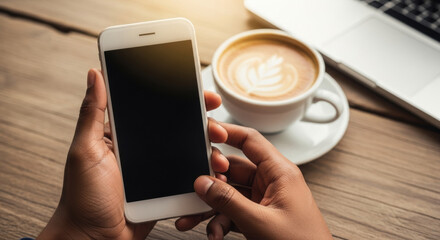 Close up of hands holding a white smartphone beside latte art coffee and a silver laptop on a wooden tabletop. Warm lighting highlights the modern technology.
