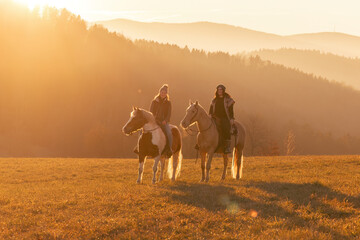 Two women riding horses for leisure in winter landscape, warm sunset light, relaxed rural atmosphere