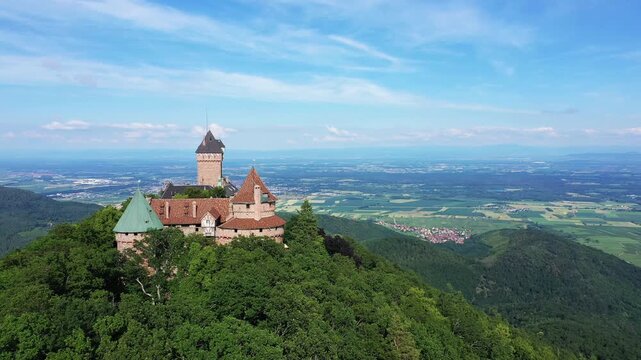 Aerial view of Chateau du Haut Koenigsbourg perched atop a lush green hill, overlooking the Alsace countryside under a clear blue sky.