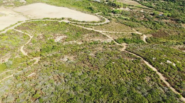 Drone shot of the rural landscape around Calenzana, featuring winding dirt roads, green fields, and scattered vegetation. The scene captures the natural beauty and open space of the Corsican country